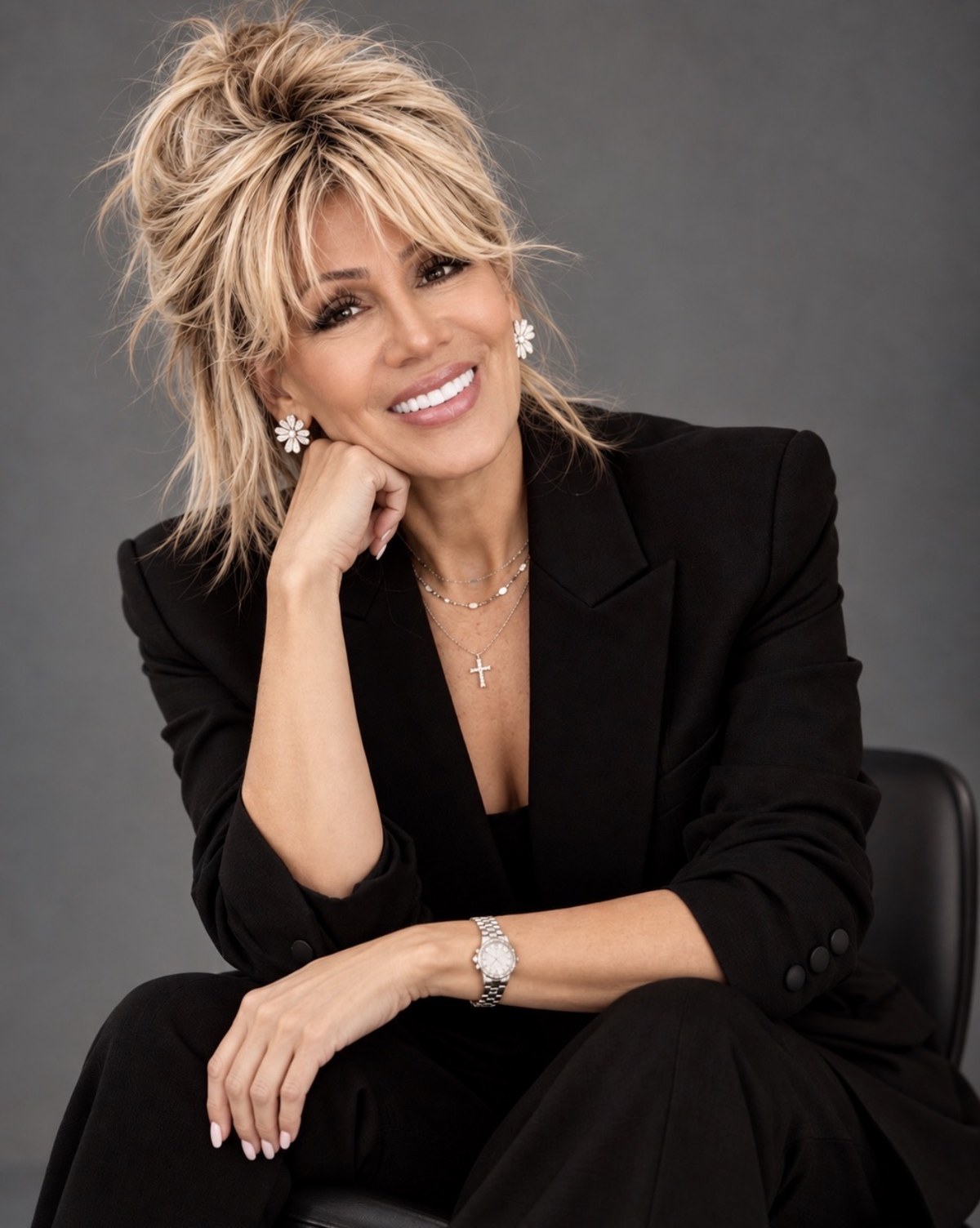 Kim Fisher in a black blazer, chin resting on her hand, smiling against a grey studio backdrop