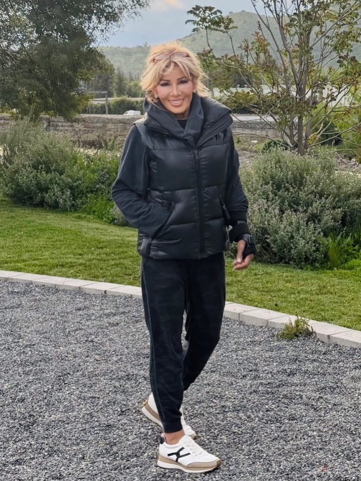 Kim Fisher walking a gravel path in a black puffer vest and joggers, green hills behind her